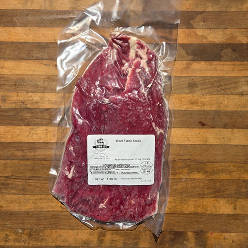A vacuum-sealed beef flank steak with a white product label sits on a wooden surface.