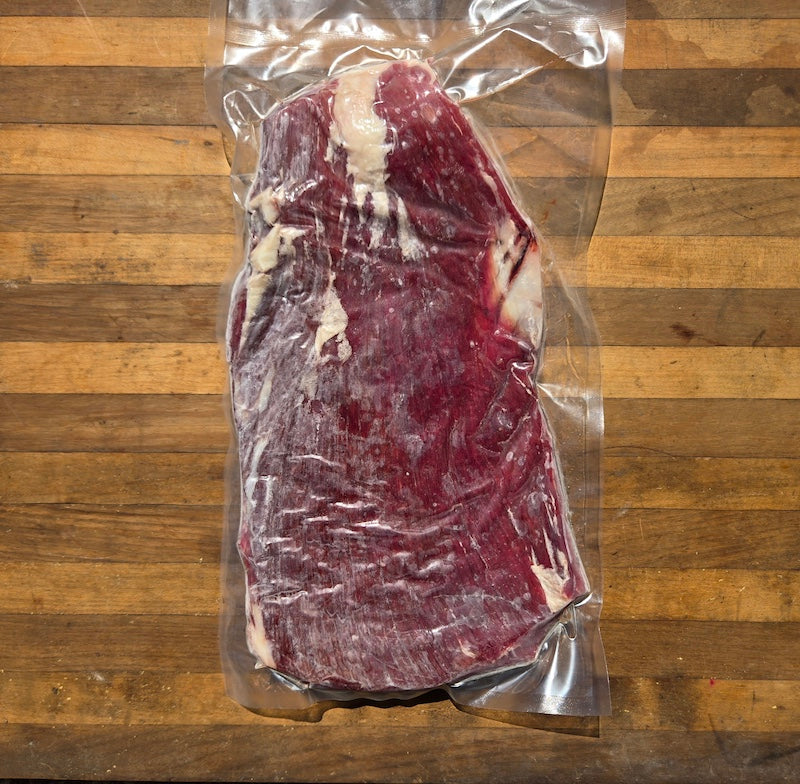 A vacuum-sealed beef flank steak sits on a wooden surface.