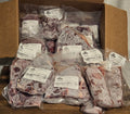 A cardboard box filled with multiple vacuum-sealed packages of raw meat, photographed on a tabletop with a tablecloth visible around it. Each package has a white label, and the meat appears frozen or covered in frost.