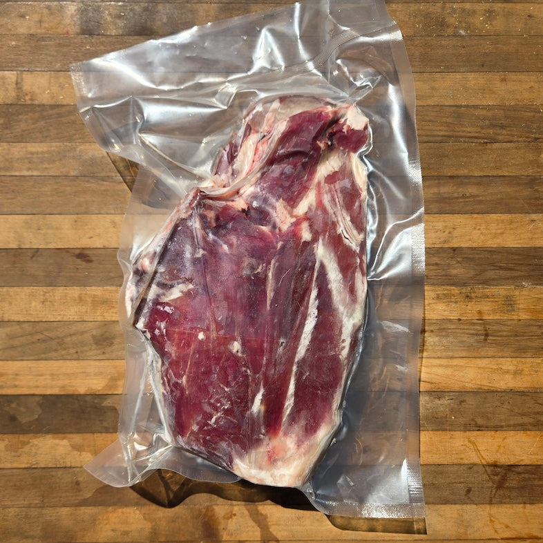 A vacuum-sealed whole lamb leg cut of meat with a white product label, placed on a wooden surface