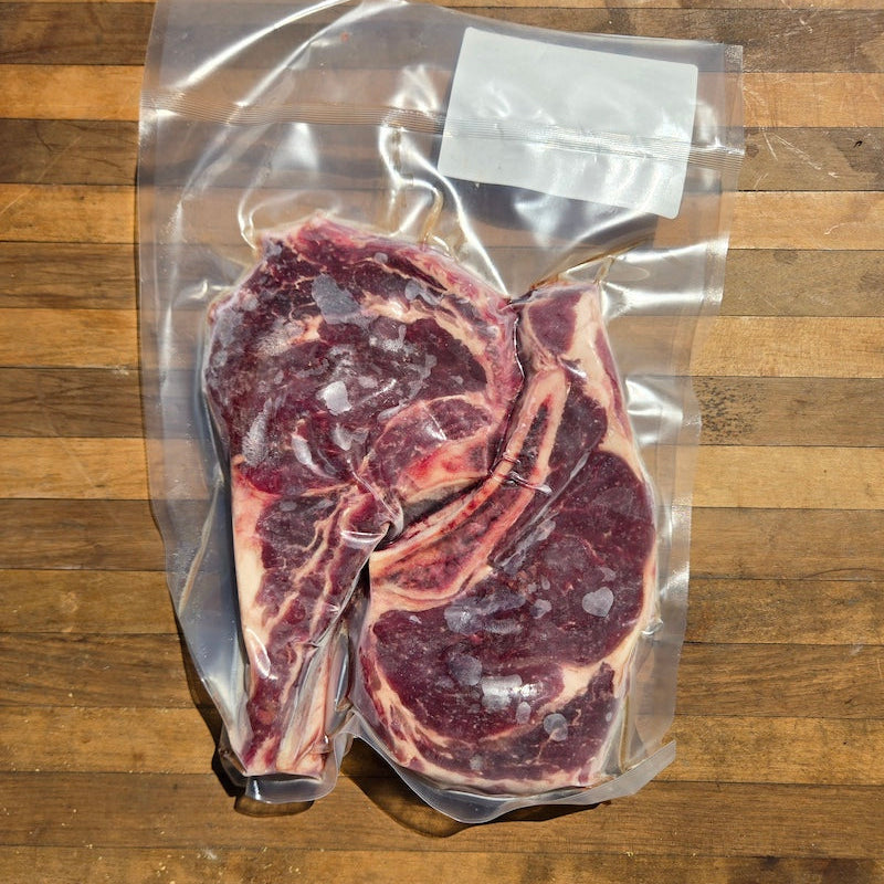 A vacuum-sealed Beef Ribeye Steak package placed on a wooden surface. The clear bag contains a thick cut of raw beef with visible bone and marbling.