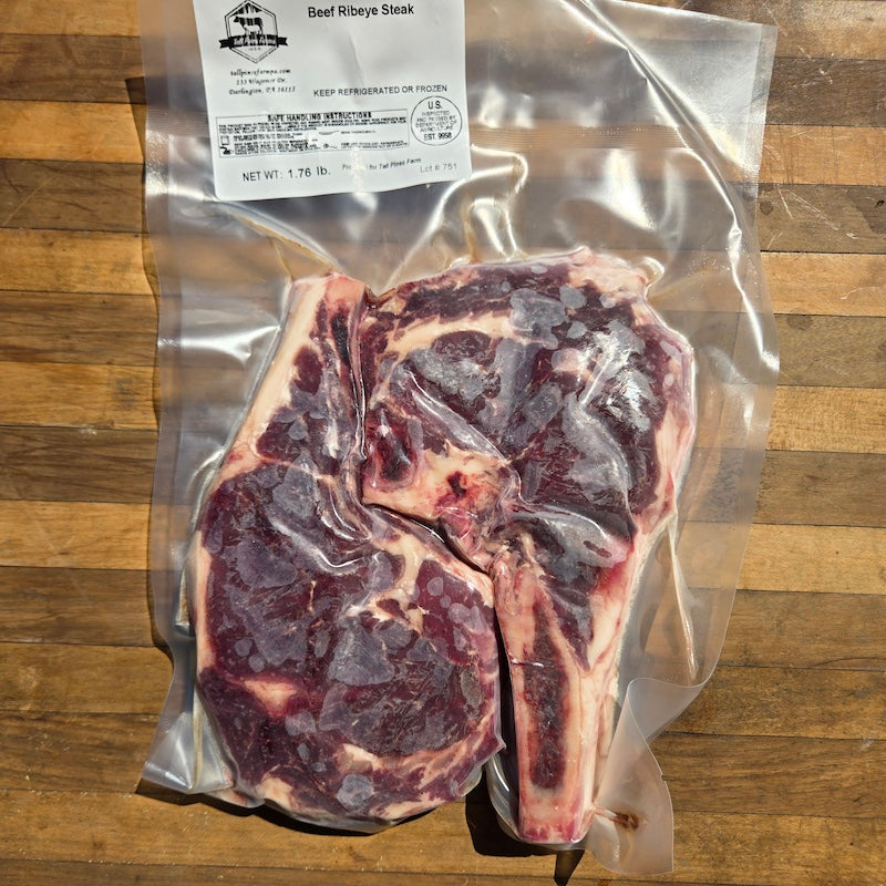 A vacuum-sealed Beef Ribeye Steak package placed on a wooden surface. The clear bag contains a thick cut of raw beef with visible bone and marbling, and a white product label is attached at the top.