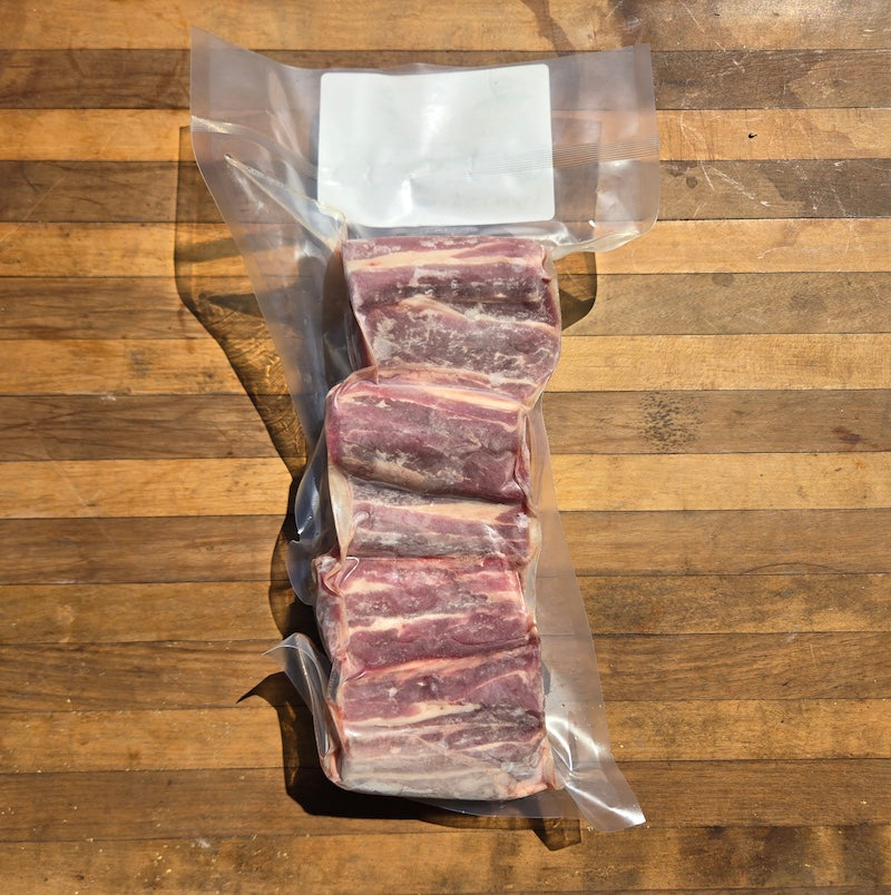 Four raw beef short ribs in a clear vacuum-sealed bag with a blank white label, placed on a wooden cutting board.