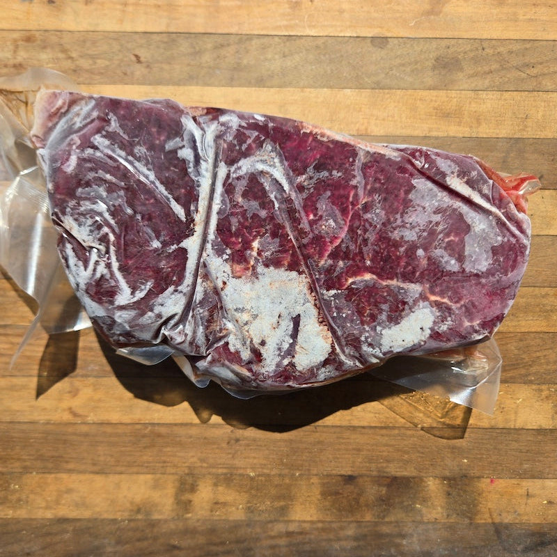 A raw, frozen cut of beef in clear plastic packaging, placed on a wooden cutting board.